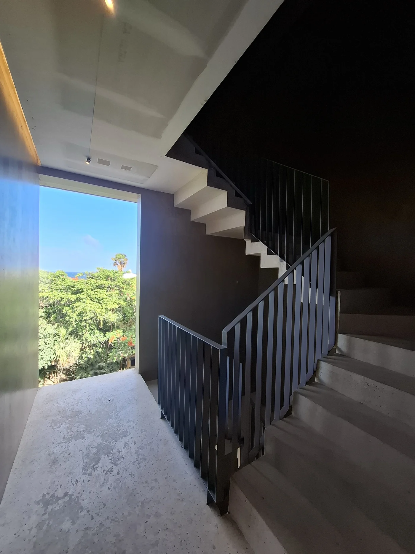 Interior stairwell of a building under construction, with an open doorway framing jungle and the sea beyond