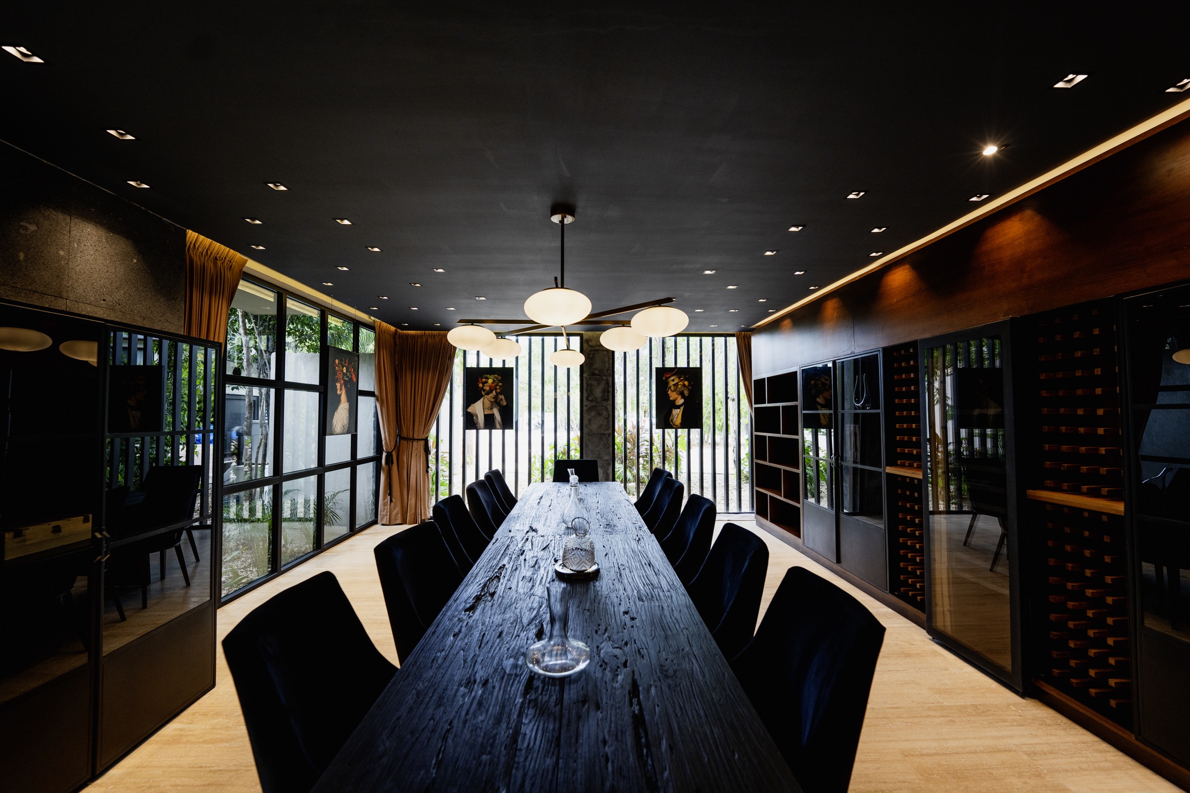 Photograph of a long charred timber table with dark blue velvet chairs under a black ceiling, wine racks behind glass along one wall and ochre curtains at the windows