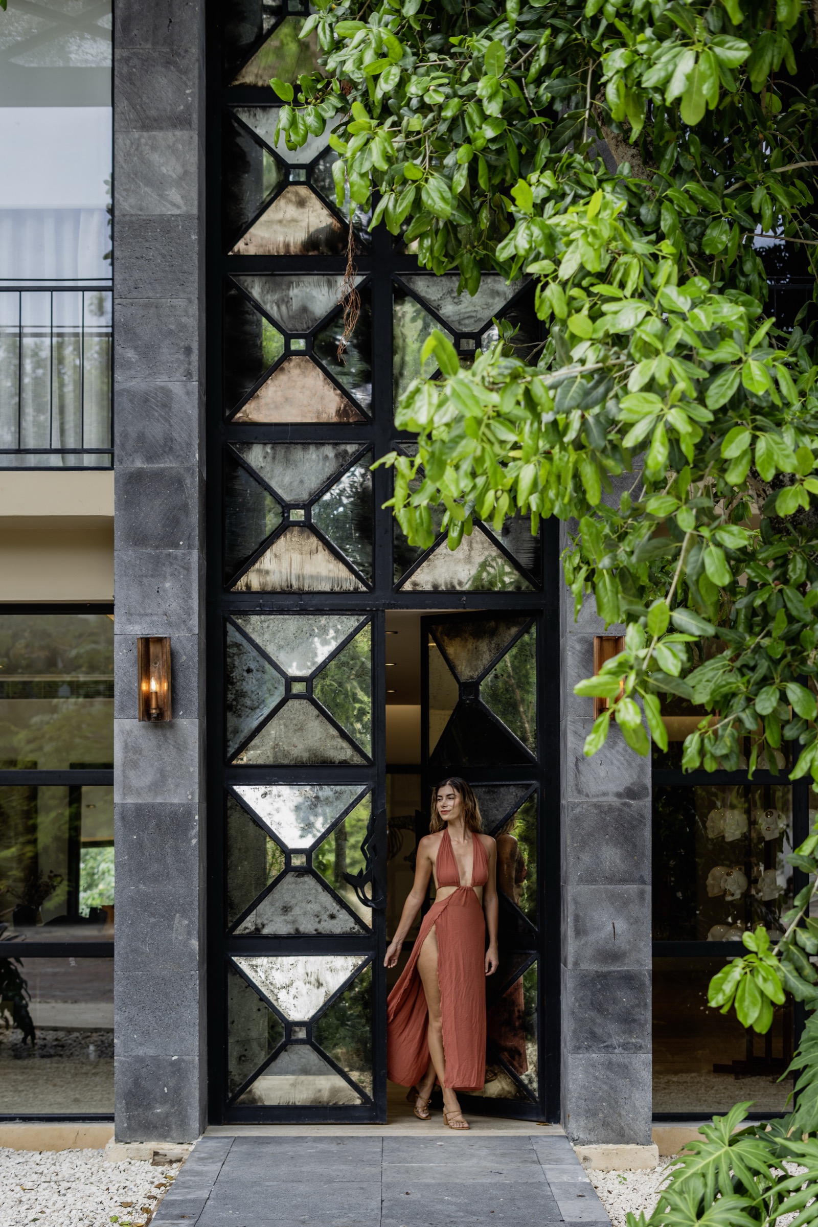 Photograph of the building entrance at Arthouse in Aldea Zamá, Tulum: a tall steel door with triangular antiqued-mirror panels set between dark stone piers, foliage overhead and a woman in a rust dress standing in the opening