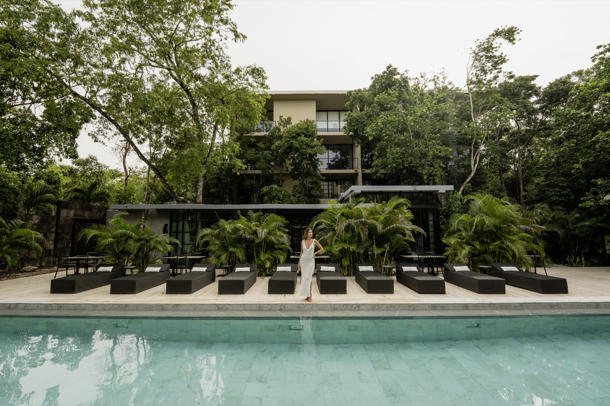 Photograph of the building entrance at Arthouse in Aldea Zamá, Tulum: a tall steel door with triangular antiqued-mirror panels set between dark stone piers, foliage overhead and a woman in a rust dress standing in the opening