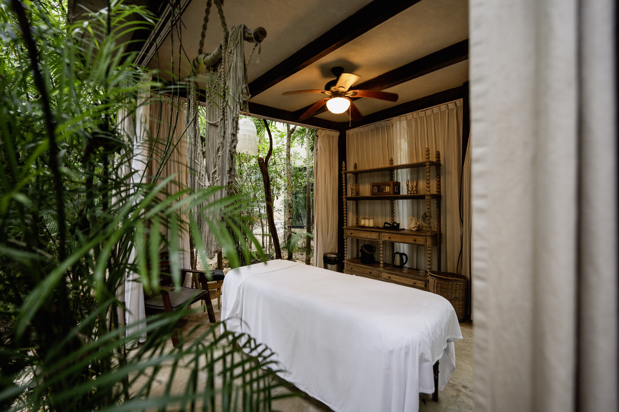 Photograph of an open-air massage cabana at Arthouse, Aldea Zamá: a linen-dressed table under a ceiling fan, a bobbin-turned wooden shelf and jungle at its edge