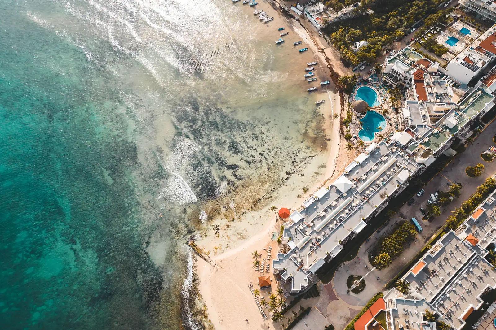 Overhead aerial view of hotel and condominium blocks with pools lining a sandy beach beside a shallow reef in Playa del Carmen