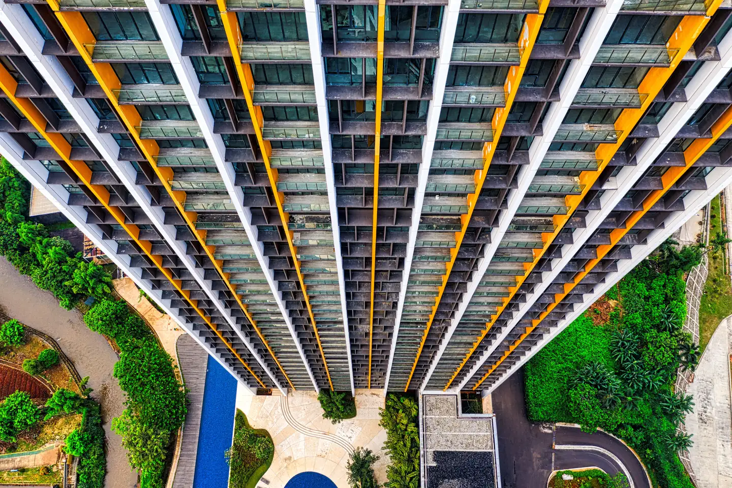 Aerial view looking straight down the side of a modern residential tower, rows of balconies stacked floor after floor above a pool and garden