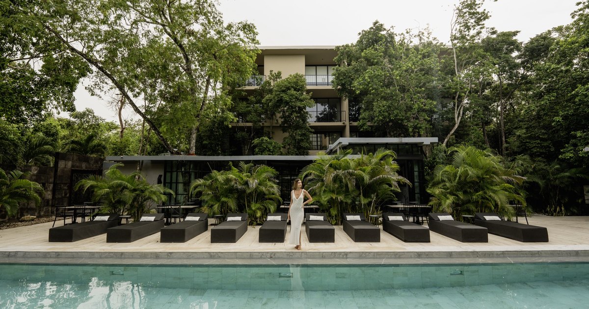 Photograph of the building entrance at Arthouse in Aldea Zamá, Tulum: a tall steel door with triangular antiqued-mirror panels set between dark stone piers, foliage overhead and a woman in a rust dress standing in the opening